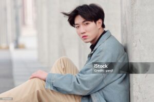 Portrait of a handsome Chinese young man in jeans sitting on ground and looking at camera.