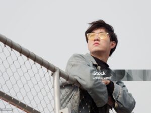 Portrait of a handsome Chinese young man standing and looking at camera with arms leaning on rail in windy sunny day, has nothing to do just waiting.